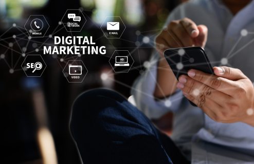 Grafika przedstawiająca siedzącego człowieka, przeglądającego telefon, z którego wychodzi napis "digital marketing" oraz ikonki reprezentujące digital marketing.