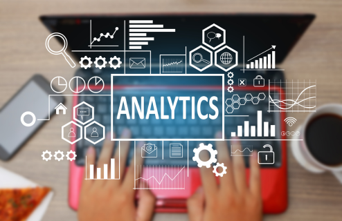 Grafika przedstawiająca analizę danych online – wykresy, ikony i napis 'Analytics' nad osobą pracującą na laptopie.